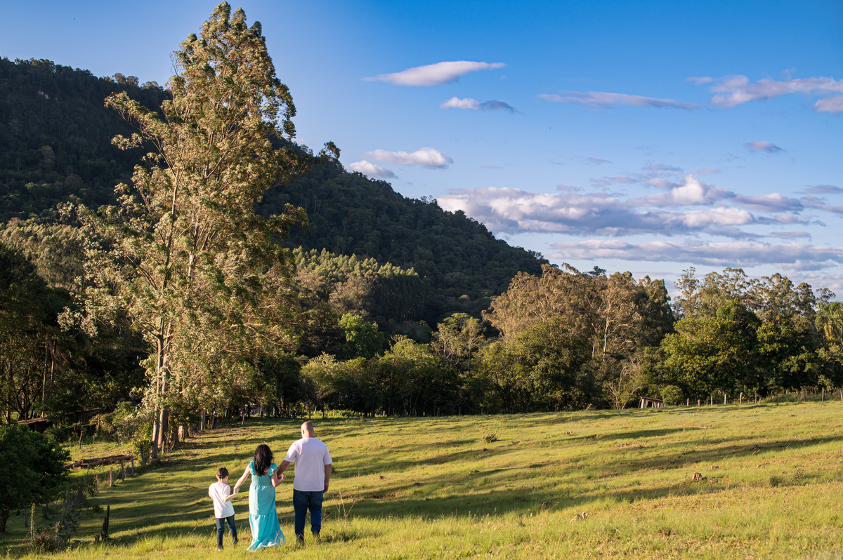 Ensaio de gestante da chácara - família caminhando, composição com o campo verde, árvores, montanha e céu azul