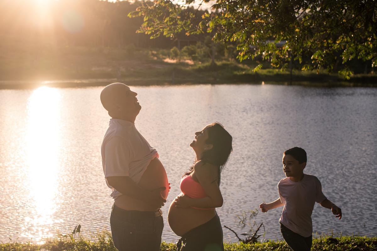 Ensaio de gestante da chácara - contra luz, por do sol, açude ao fundo, pai e mãe com a barriga de fora, criança correndo