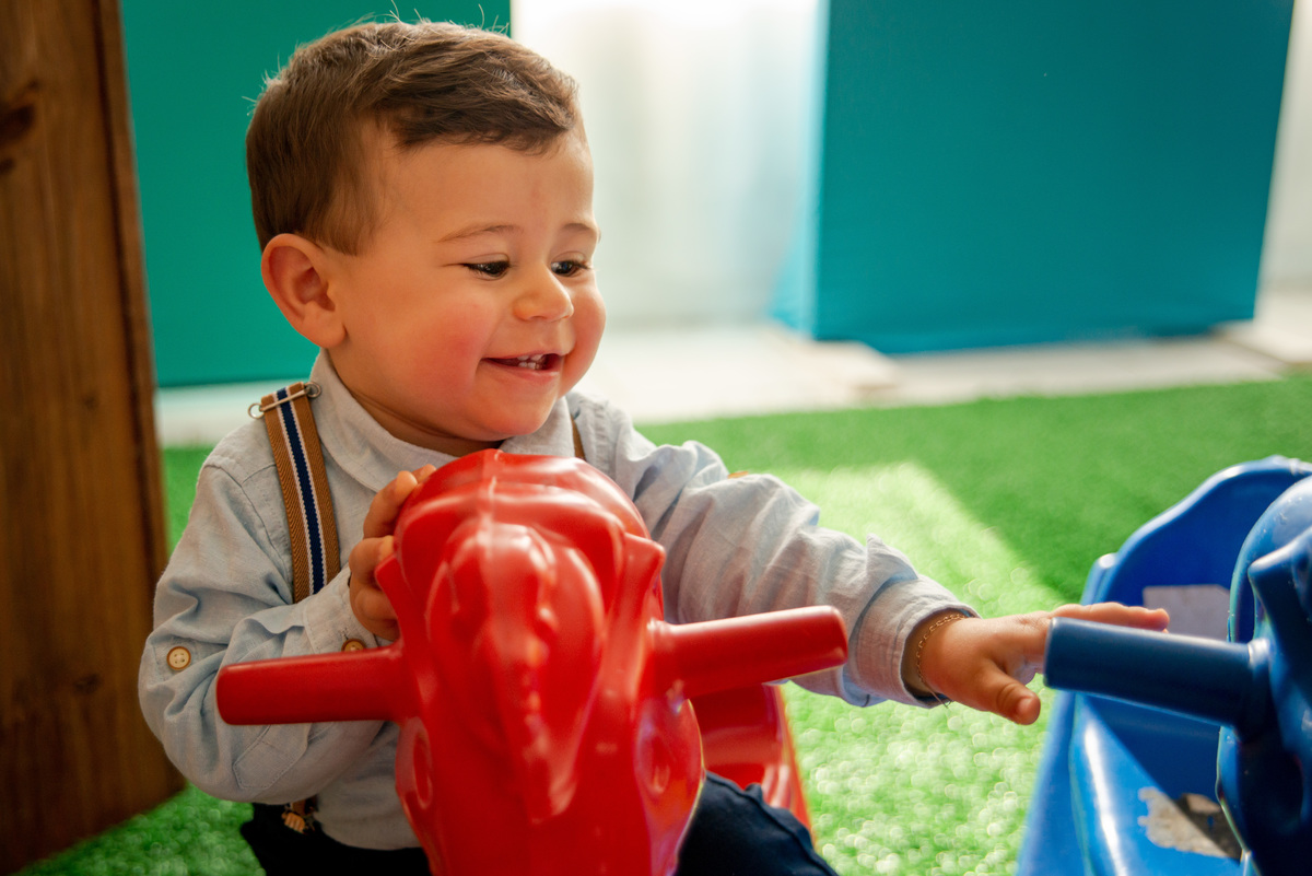Festa de aniversário de um aninho, menino brincando com 2 cavalinhos, sorrindo, mostrando os dentinhos