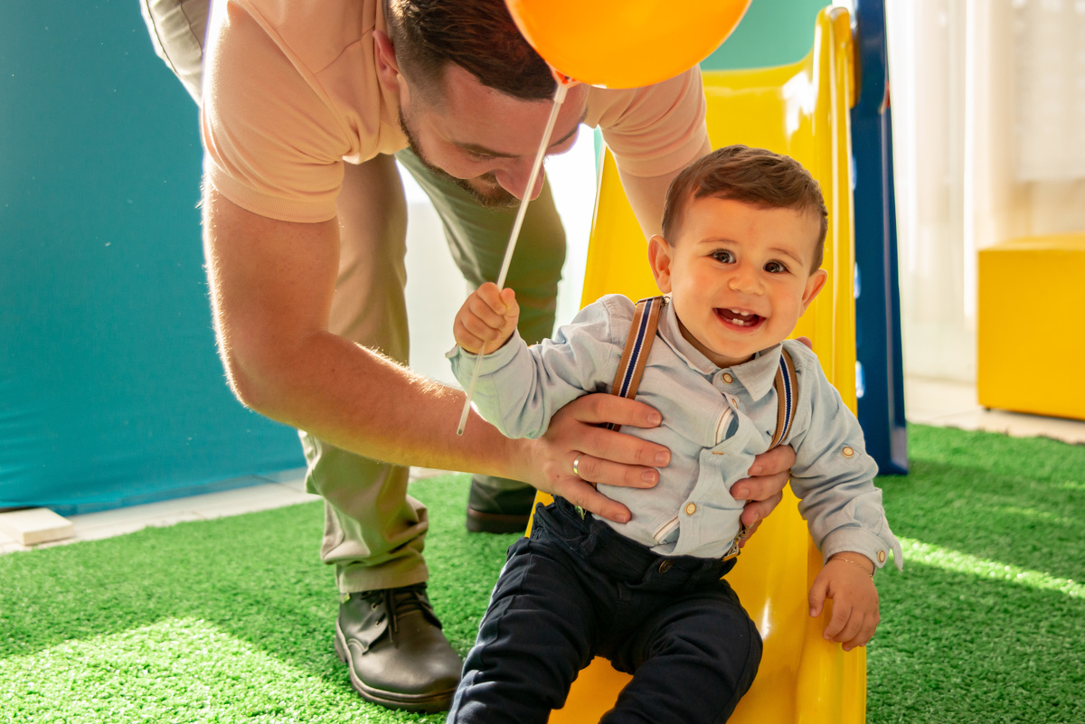 Menino na festa de um aninho brincando no escorregador com o pai, sorrindo e mostrando os dentinhos
