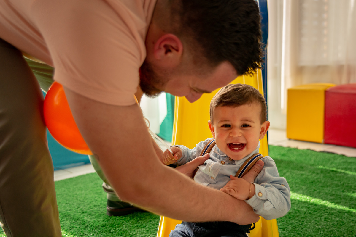 Menino na festa de um aninho brincando no escorregador com o pai, sorrindo e mostrando os dentinhos