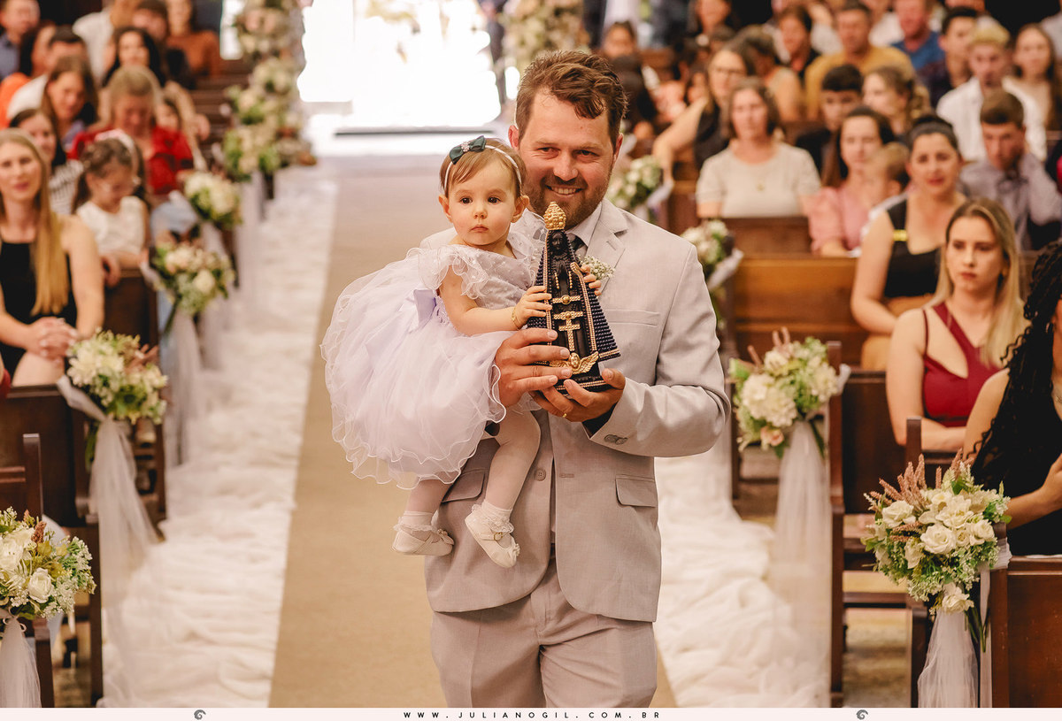 padrinho segurando a daminha e levando nas mãos a imagem de nossa senhora aparecida na entrada da cerimônia na igreja matriz em Rio Azul, Paraná