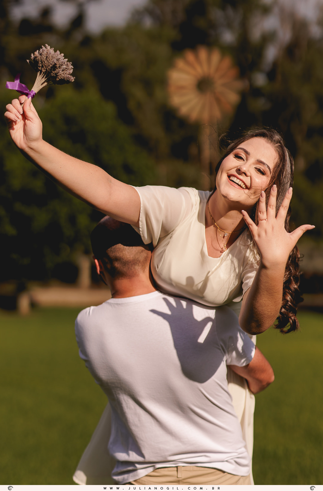 noivo segurando a noiva, ele está de costas e ela de frente pra camera sorrindo e mostrando a mão com a aliança, na outra segurando um buquê de lavanda
