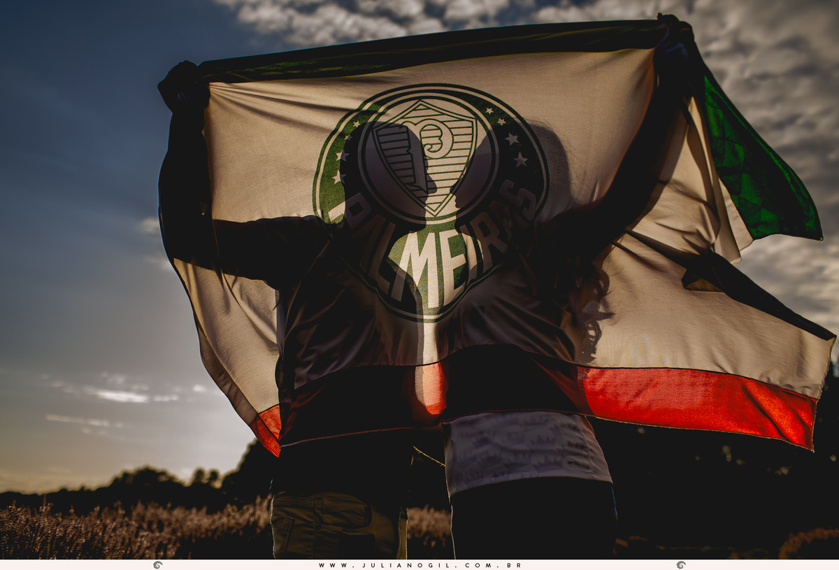 silhueta do casal no pôr do sol, eles estão segurando a bandeira do palmeiras na frente deles com efeito que o rosto do casal fica bem no meio do brasão do time