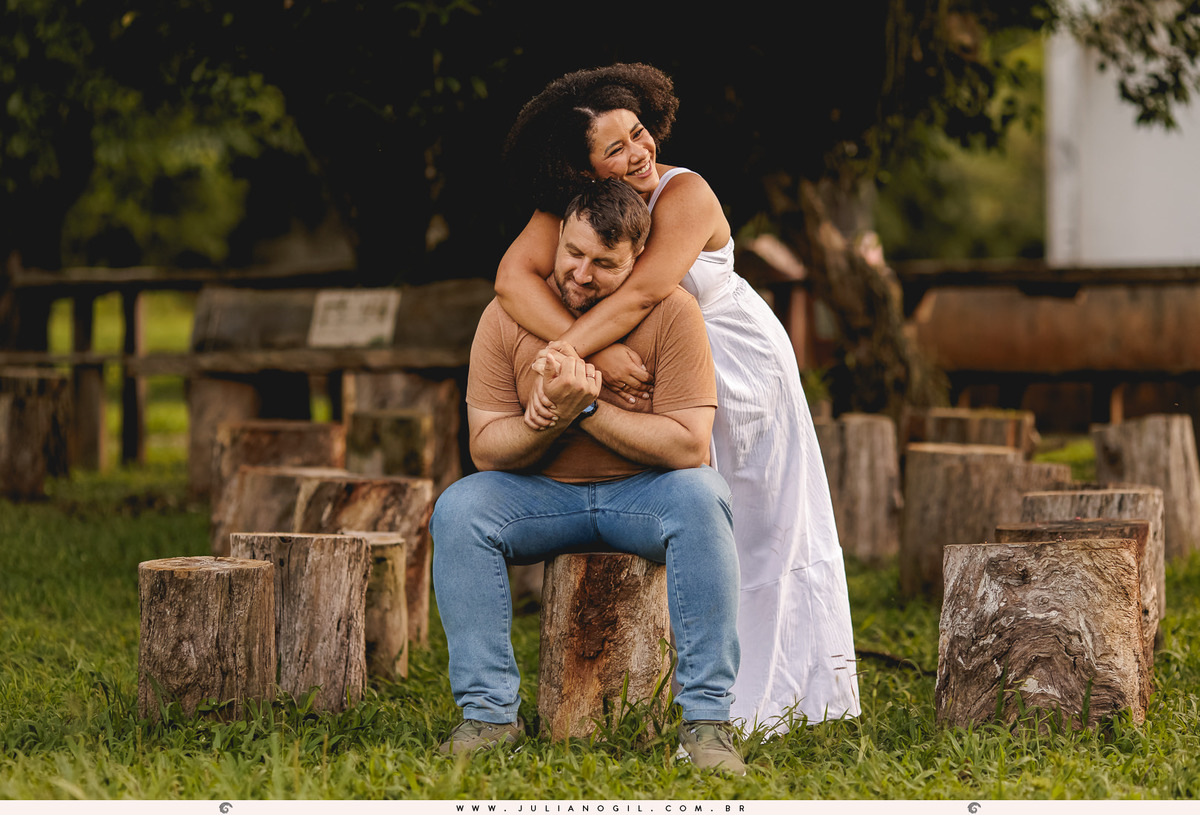 noiva abraçando o noivo pelas costas, ele está sentado em um toco de árvore