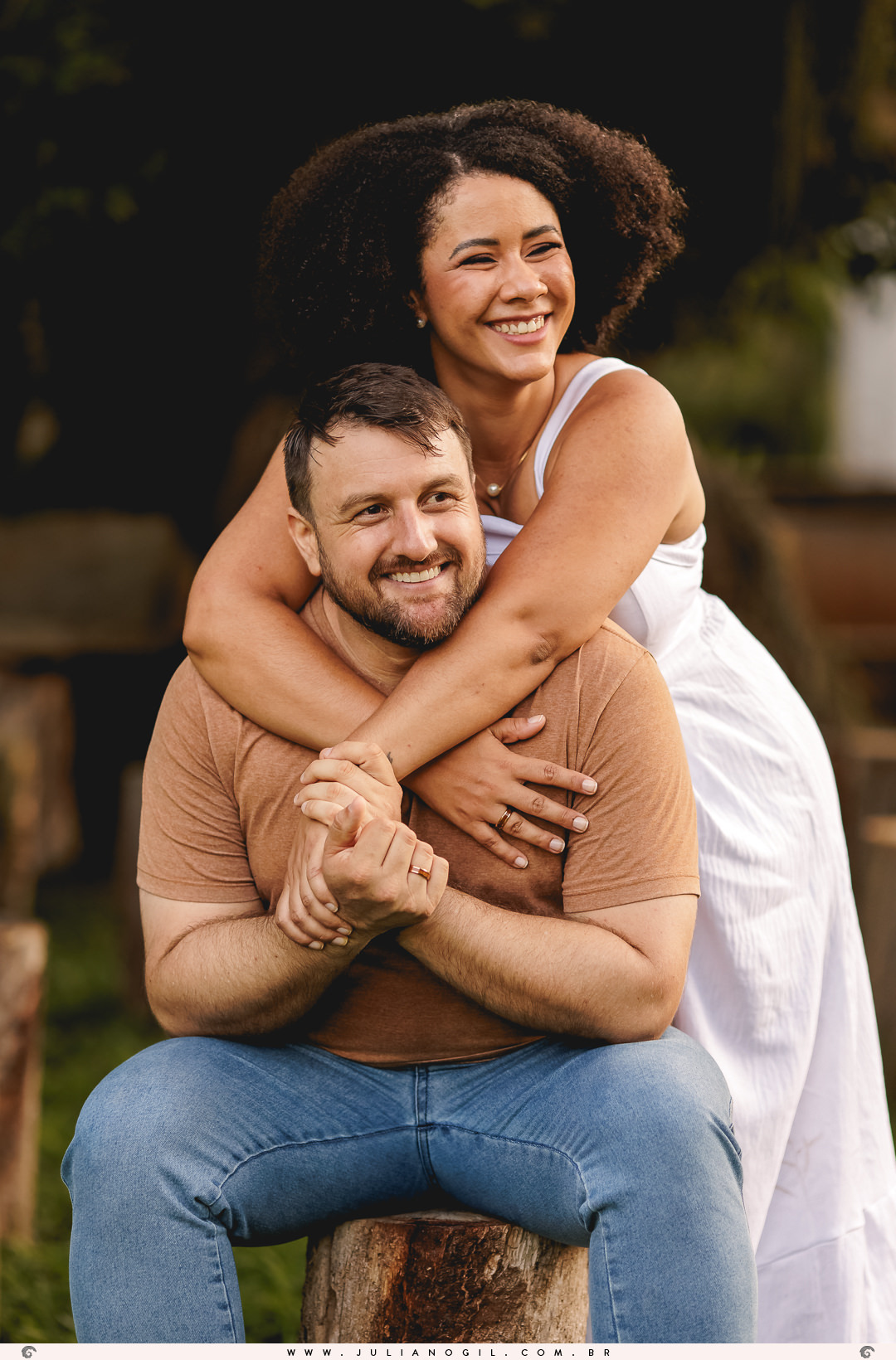 noiva abraçando o noivo pelas costas, ele está sentado em um toco de árvore