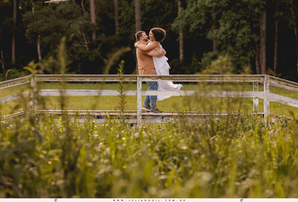casal se abraçando em uma ponte ao fundo, na frente vários arbustos