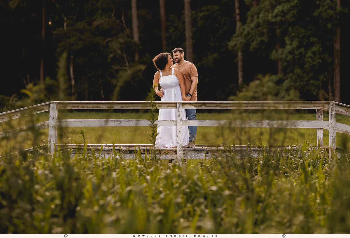 casal se abraçando em uma ponte ao fundo, na frente vários arbustos