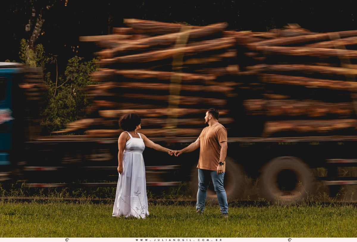 casal focado em primeiro plano e ao fundo uma carreta carregada de toras passando desfocada