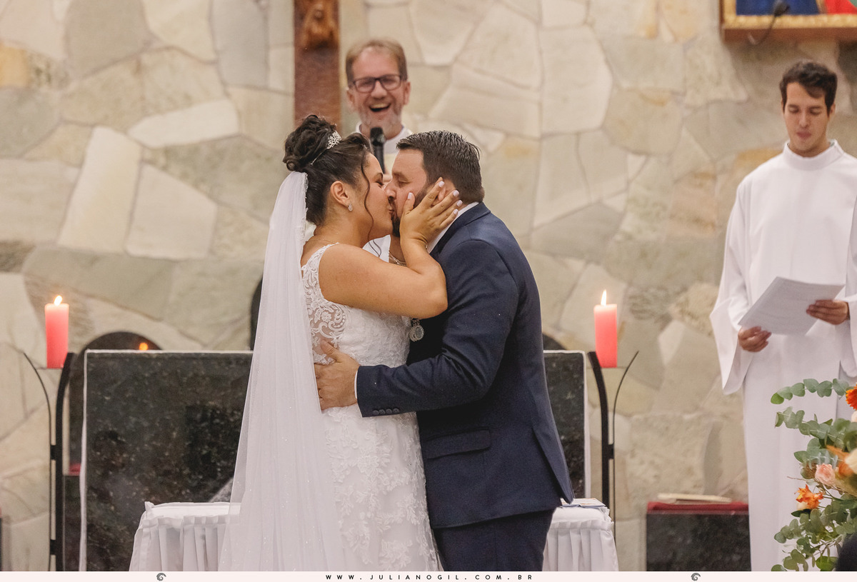 casal se beijando em primeiro plano e padre rindo ao fundo