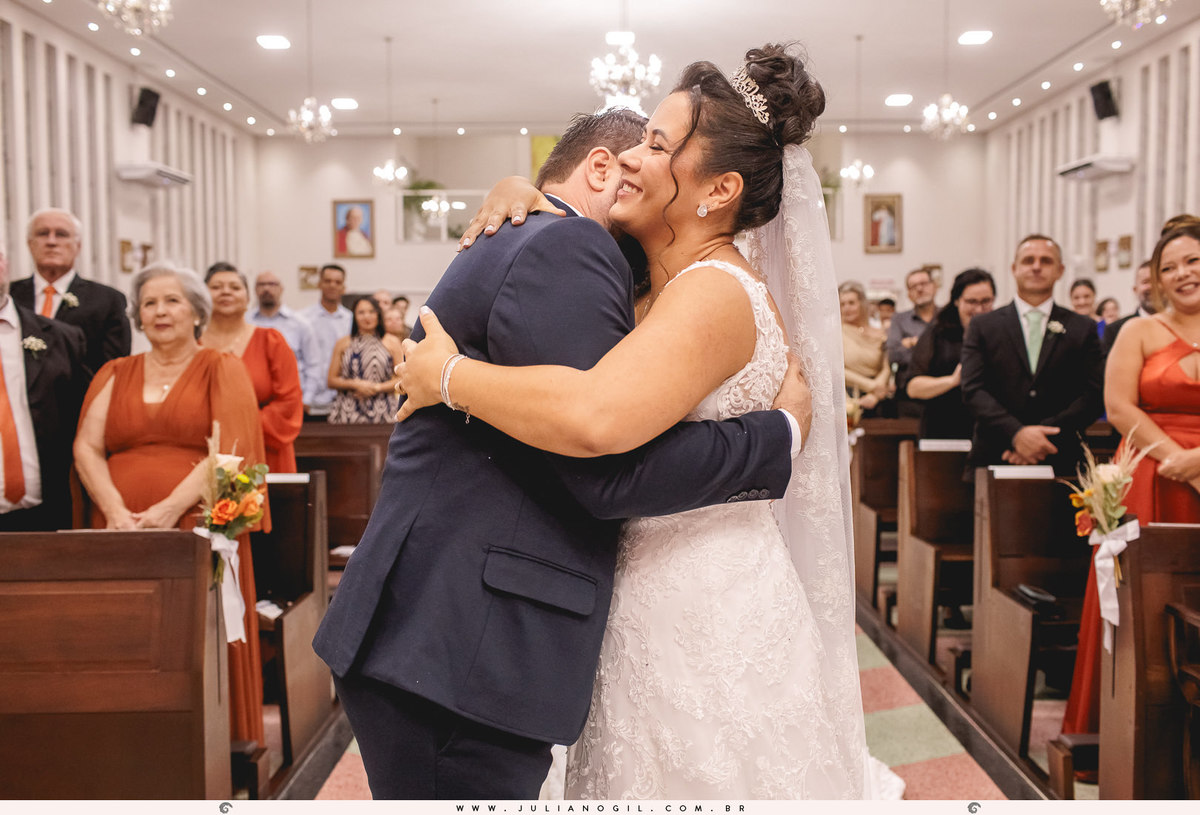 casal se braçando em frente ao altar, ao fundo convidados sorrindo e olhando
