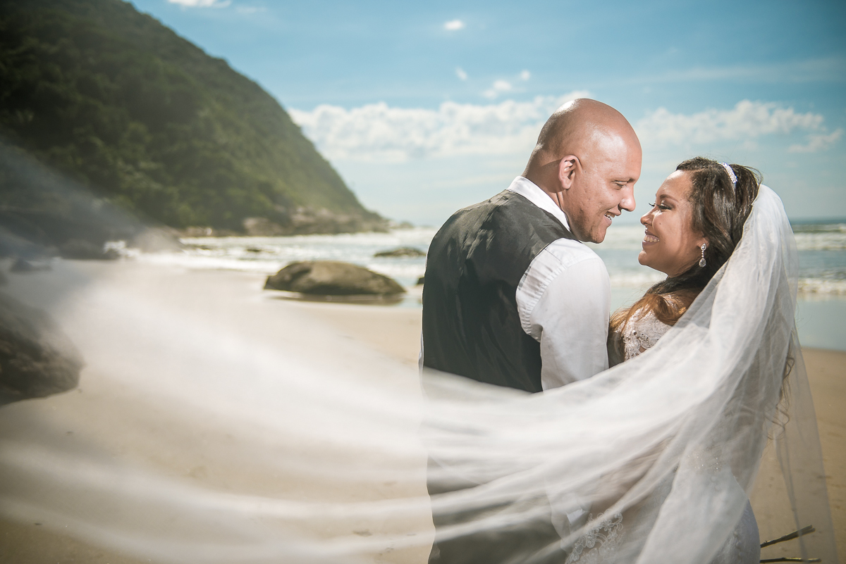 Véu da noiva ao vento com olhar apaixonado para o noivo na praia de Guarujá.