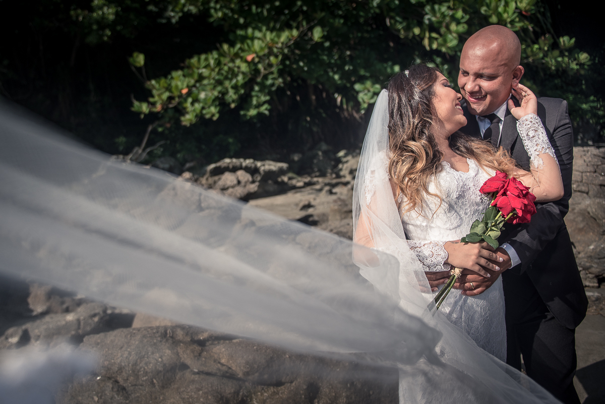 Véu da noiva leva o olhar até o casal que está sorrindo na praia