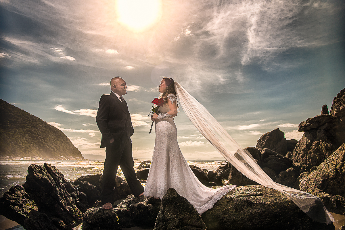 Casal em cima das pedras da praia com o sol sobre eles.
