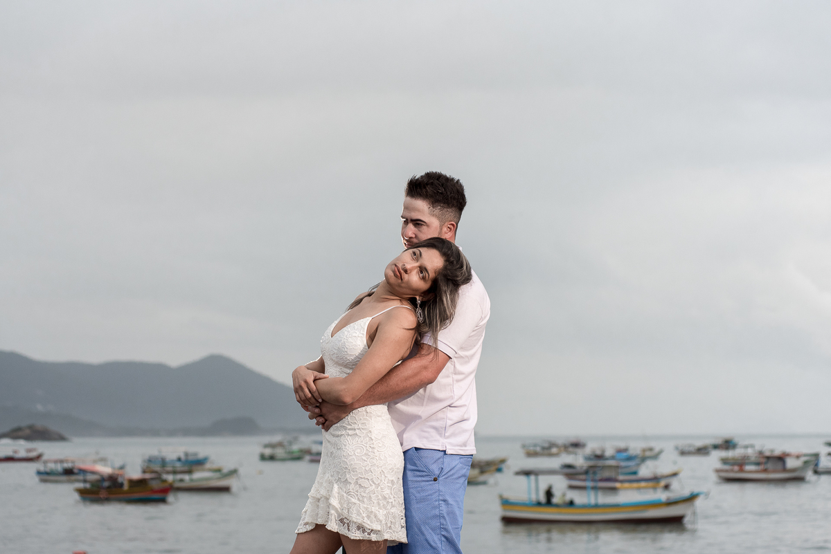O casal posa entre barcos e céu aberto para foto no ensaio pre wedding na praia.