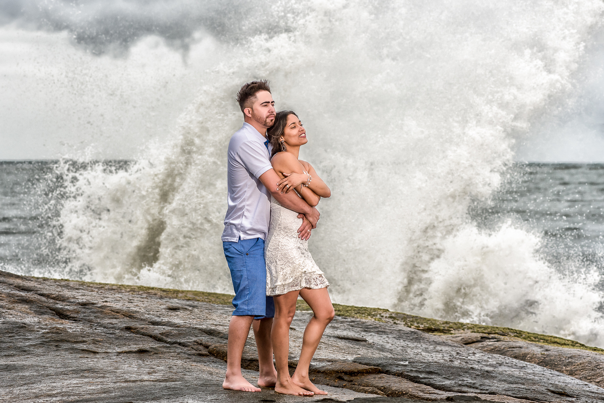 Casal abraçado quando a onda bate na pedra e forma uma onda maravilhosa, tudo isso na fotografia de ensaio de pre wedding na praia de Iporanga SP