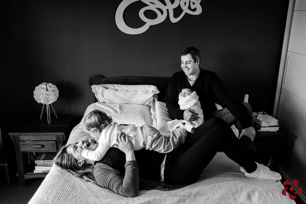 A família está no quarto, em cima da cama de casal em uma momento de interação e carinho.