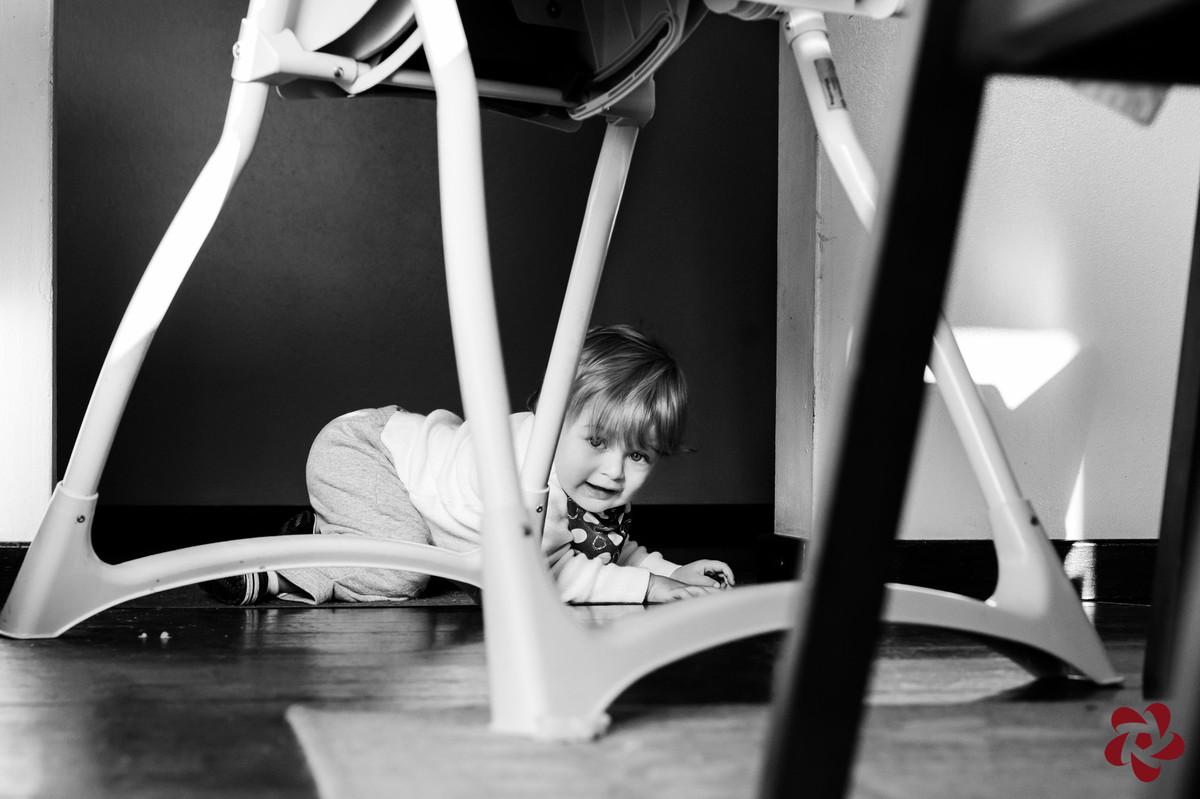 Enquanto Otávio brinca embaixo da mesa entre as cadeiras, engatinhando, ele olha para a câmera.