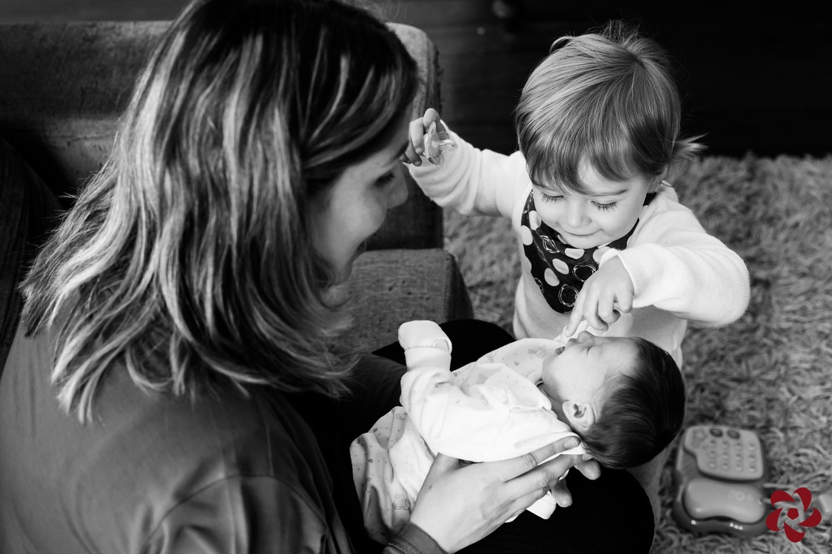 Júlia está no colo da sua mãe Natácia e Otávio, o irmão mais velho, brinca de encostar na sua boca e nariz.