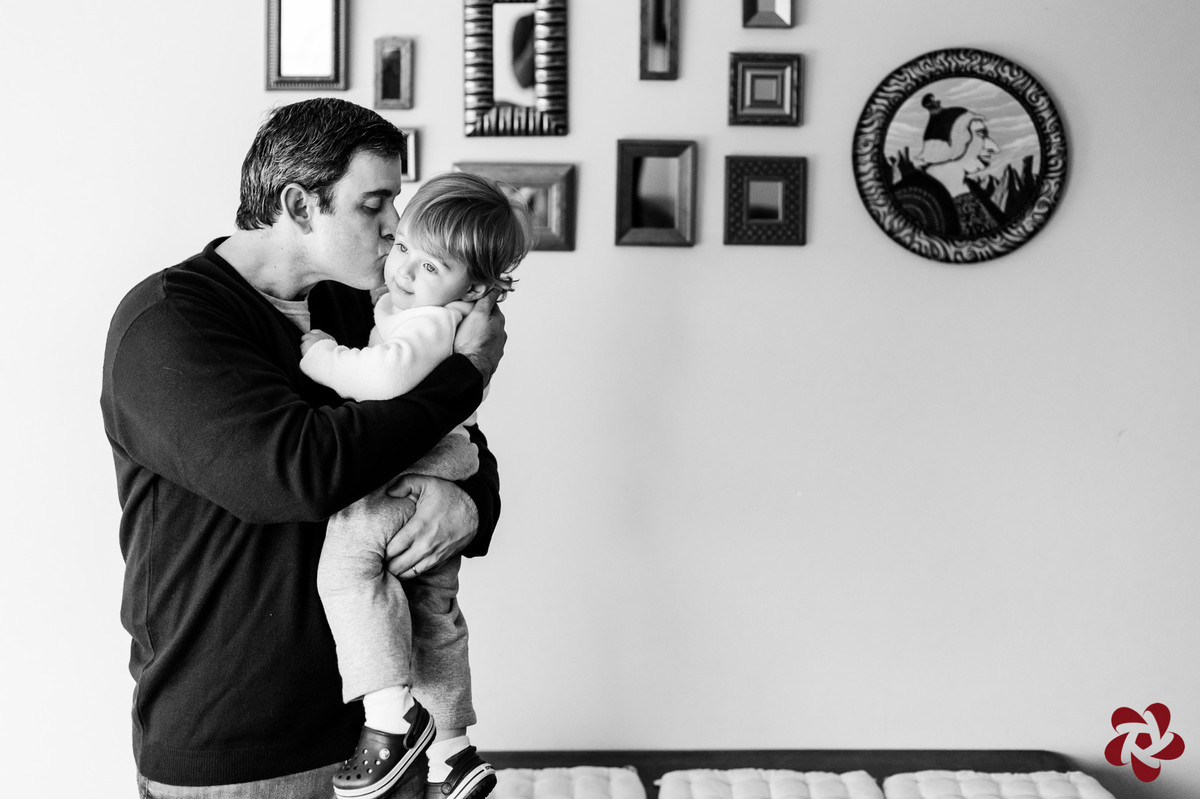 Rodrigo está com Otávio no colo, beijando seu rosto, em frente a uma parede decorada com quadros pequenos.