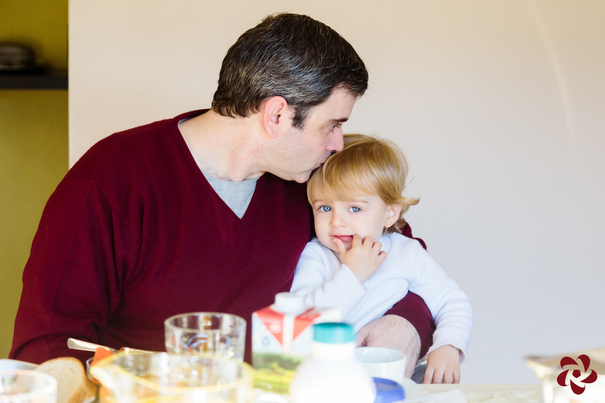 Otávio está sentado no colo do pai na mesa do café, olhando para a câmera com seus olhos azuis enquanto o pai lhe dá um beijo na cabeça.