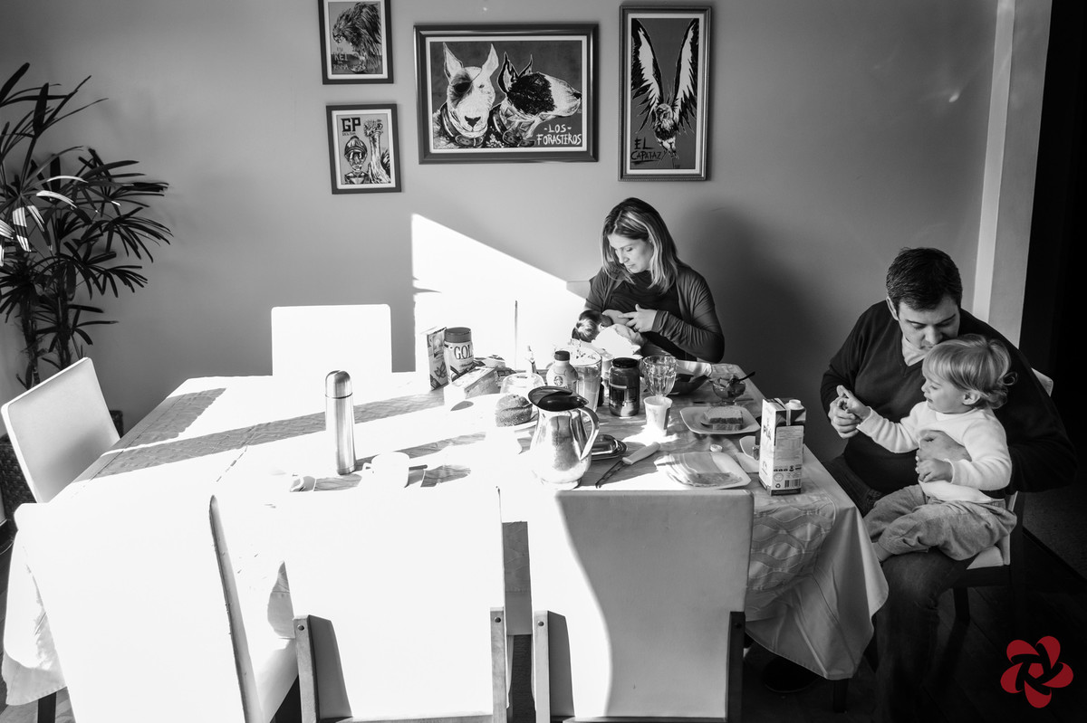 A família está reunida na mesa do café da manhã, a mãe está amamentando a filha e o pai está dando atenção ao filho mais velho