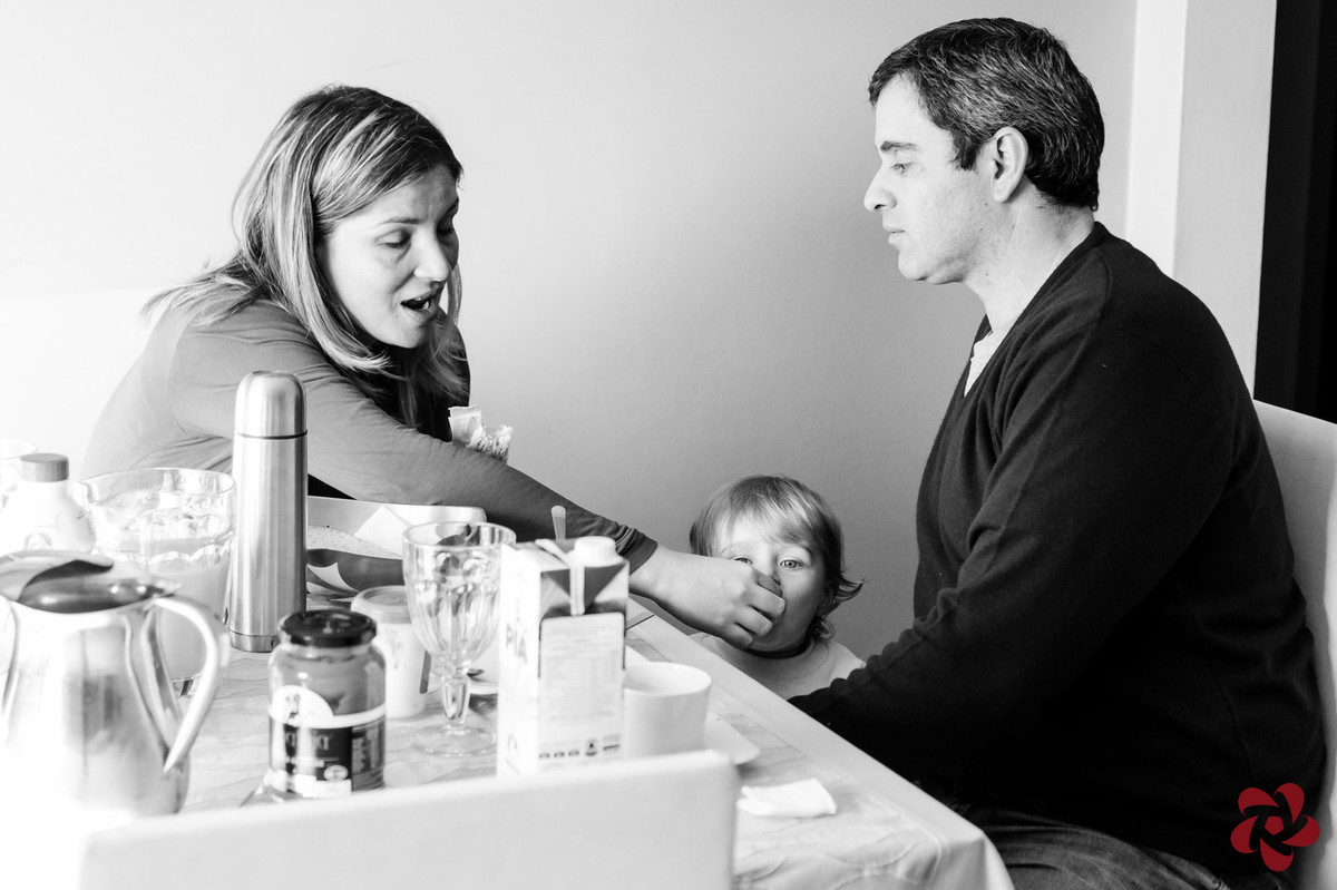 A mãe está dando pão na boca do filho que está parado ao lado do pai, na mesa do café da manhã de domingo.