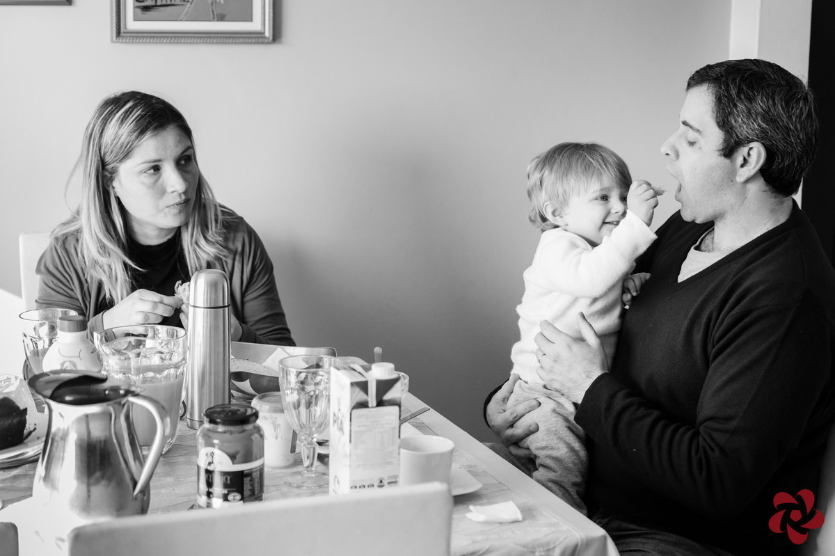 Otávio senta no colo do pai durante o café da manhã e lhe dá um pão na boca enquanto a mãe observa.