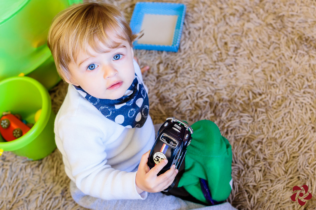 Otávio está sentando no tapeta da sala brincando com seus carrinhos e está olhando para a câmera com seus lindos olhos azuis.