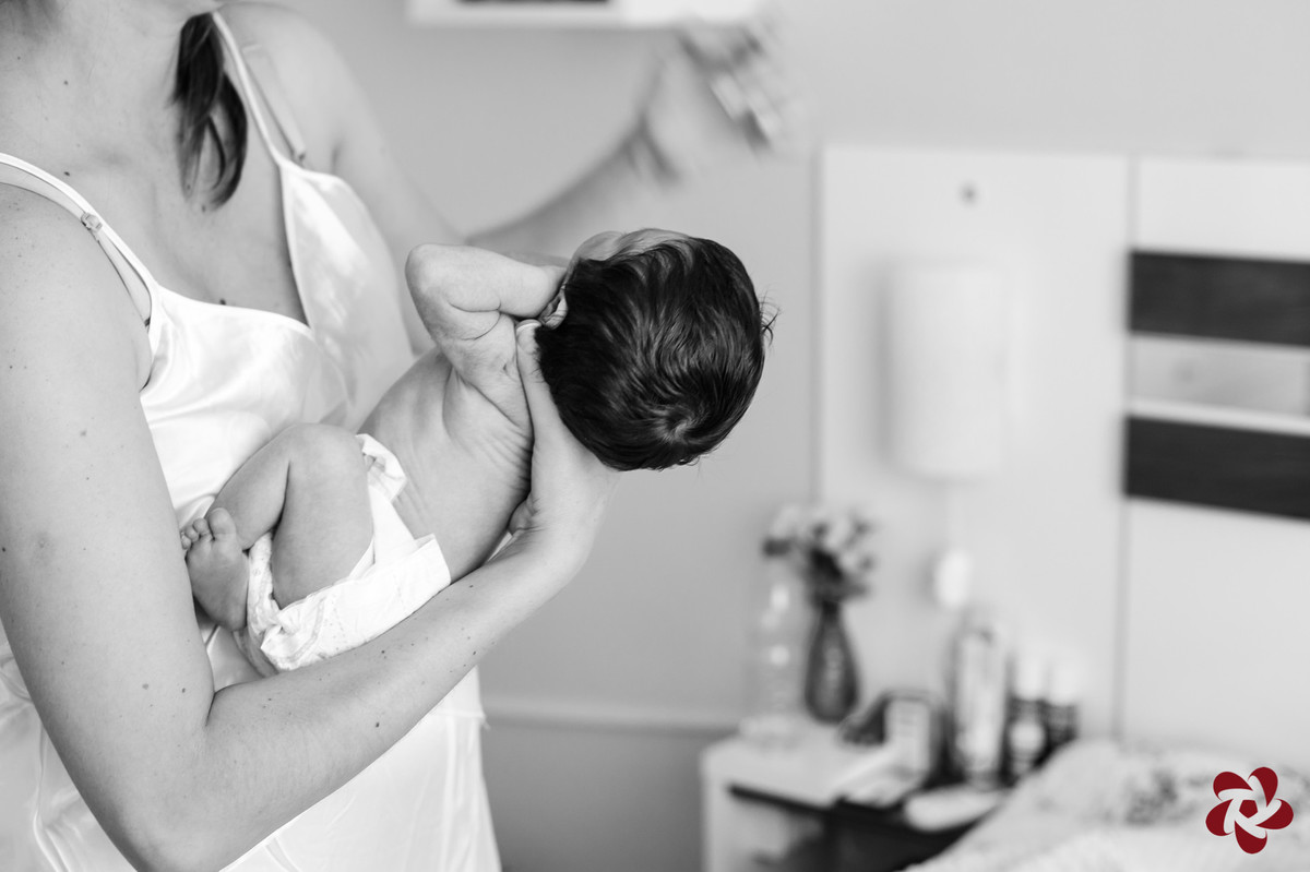 Foto em preto e branco da mamãe Kátia no quarto segurando o bebê no colo com a mão direita e apoiando no antebraço o corpinho