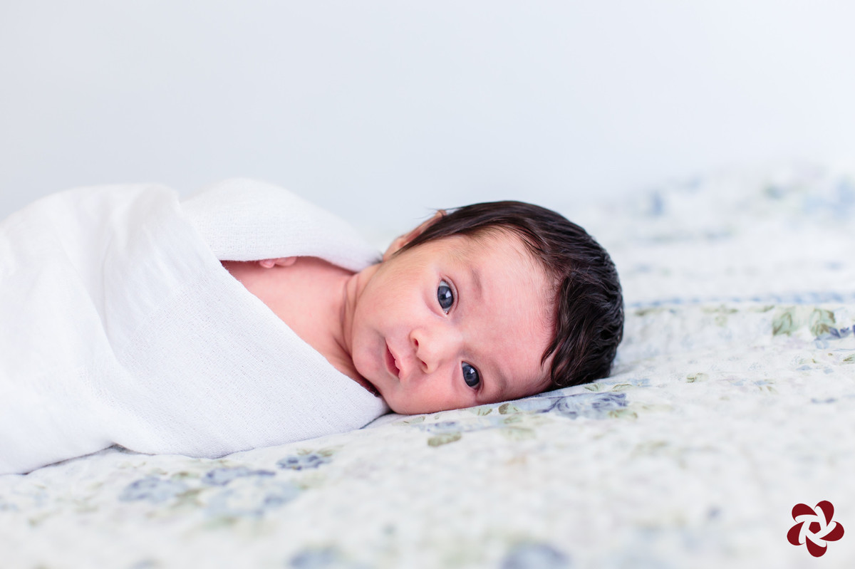 Henrique deitando na cama com colcha branca flores pequenas azuis e folhas verdes. Ele está acordado. de lado, enrolado numa fralda branca e olha atentamente para a câmera fotográfica.