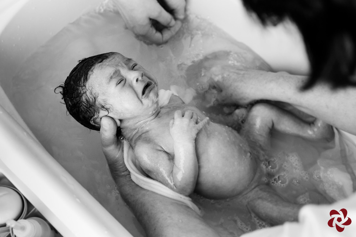 Foto em preto e branco. Henrique chora durante o banho dentro da banheira enquanto sua avó e sua mãe dão banho.
