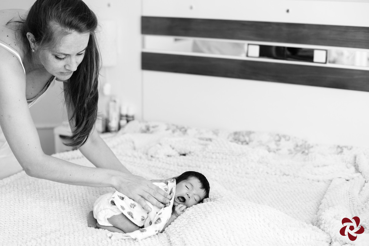 Foto em preto e branco da mamãe Katia acalmando o Henrique que está deitando na cama de casal de bruços, joelhos flexionados e tapado com uma fralda de pano