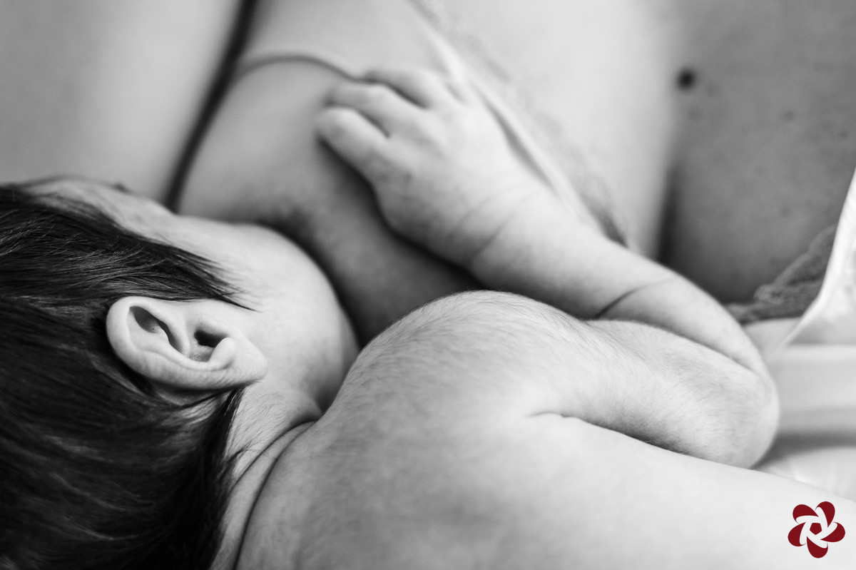 Foto em preto e branco, imagem com close no seio da mãe amamentando o filho, destacando os pelos das costas e braços e cabelo do bebê que está com a mão apoiada no seio