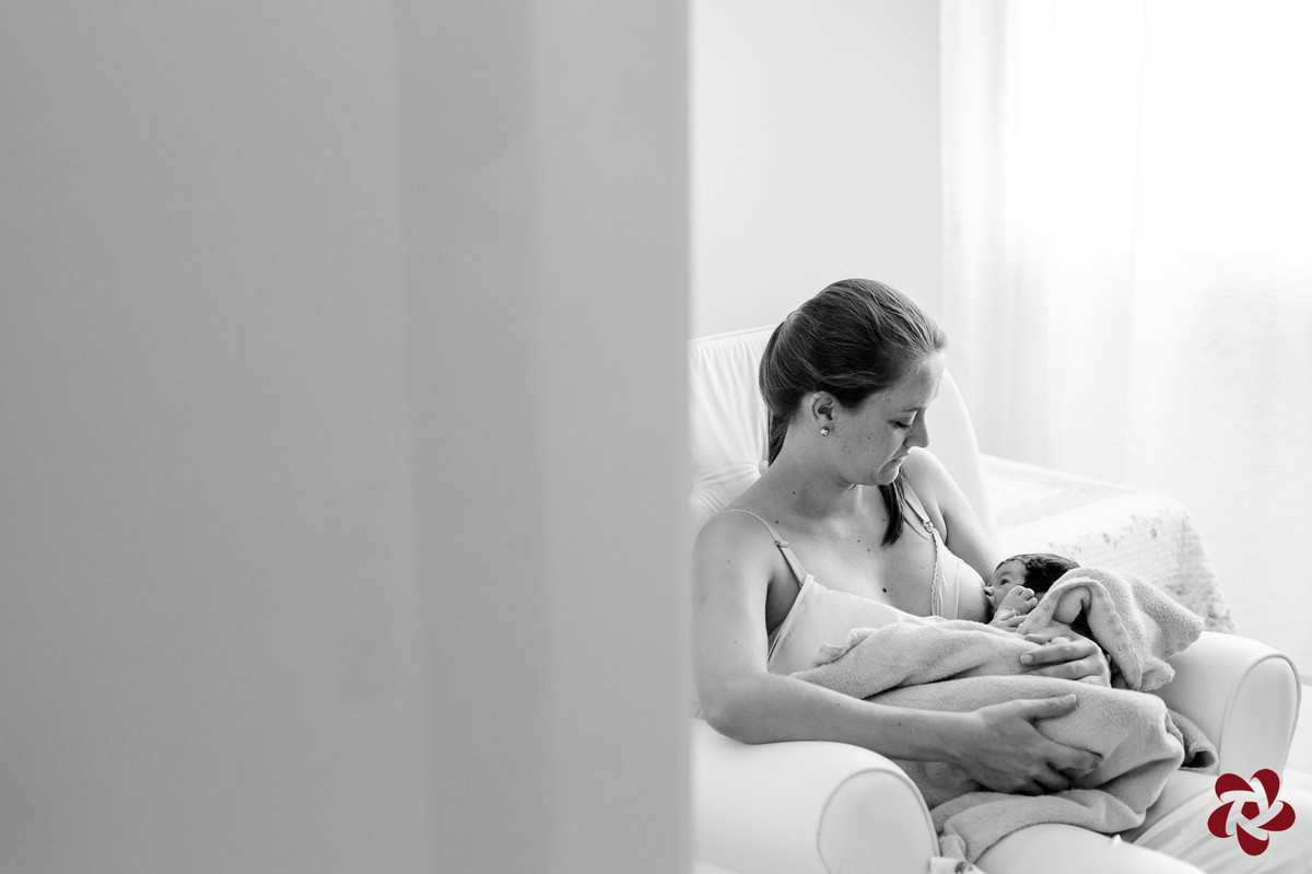 Foto em preto e branco. Katia está está no quarto sentada em uma poltrona, amamentando seu filho Henrique, que está enrolado em um cobertor, iluminados com suave luz vinda da janela