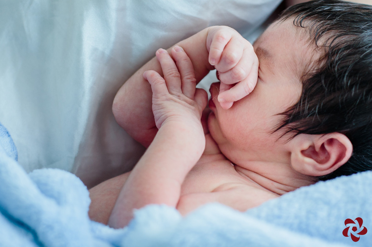Bebê Henrique deitado com a mão direita sobre os olhos e a mão esquerda apoiada no antebraço direito, enrolado em um cobertor azul claro