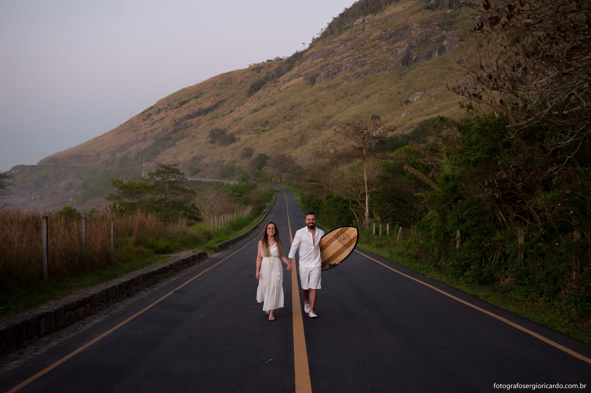Noivos caminhando na estrada de mãos dadas junto com uma prancha de surfe na praia do secreto.