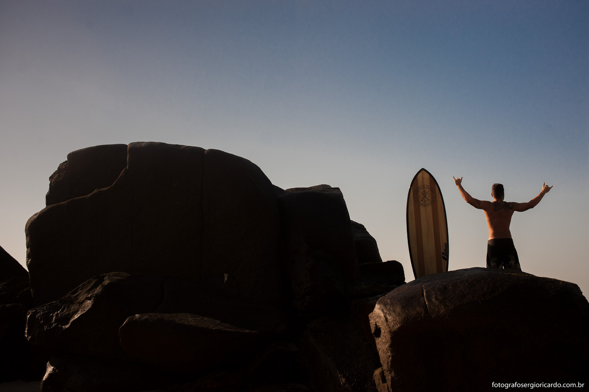 Retrato do noivo com sua prancha de surfe em cima das pedras na praia do Secreto.