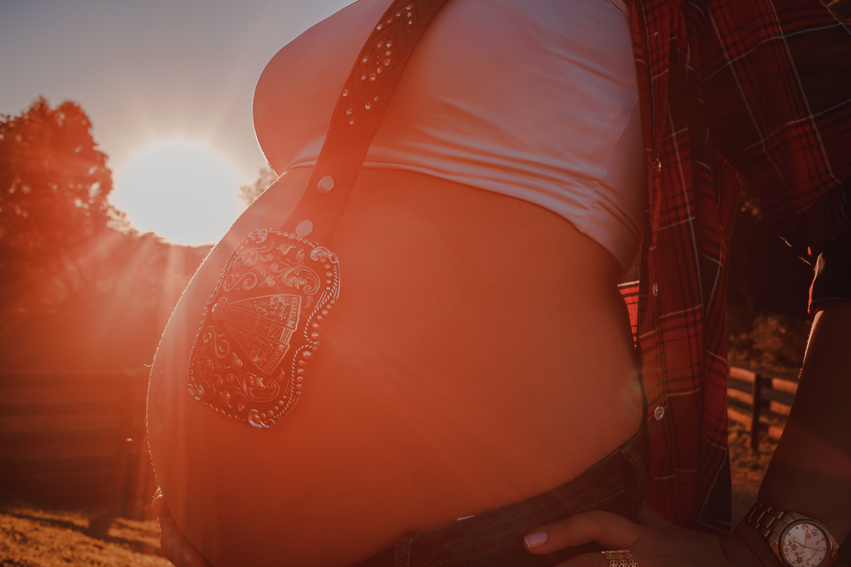close de foto de barriga de gestante com os raios do sol ao fundo refletindo na fivela que ela traz de nossa senhora aparecida