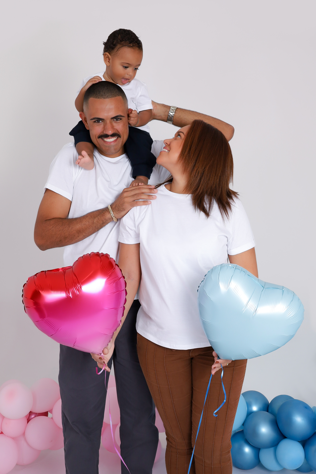 Imagem de uma gestante sorridente em meio à natureza, segurando balões azuis e rosa durante seu ensaio de revelação. Ao lado, o parceiro celebra o momento com alegria. A cena transmite emoção, surpresa e conexão familiar, registrando o instante em que des