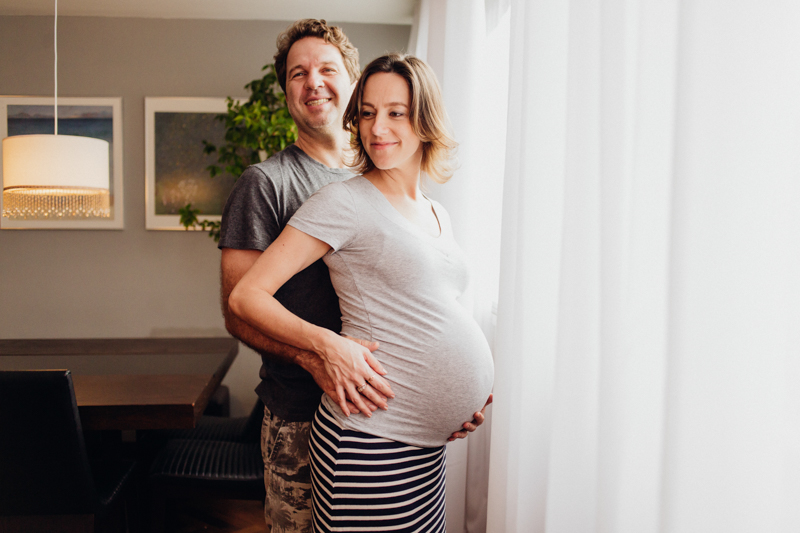 Foto do Evandro e da Tetê no apartamento onde vivem. Foto do casal esperando seu primeiro filho. Foto do casal na janela feita com luz suave. Uma foto leve e espontânea. Fotografia de família, ensaio gestante, fotos de grávidas. 