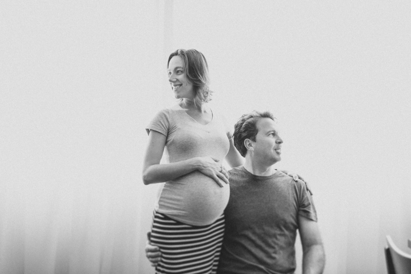 Foto do Evandro e da Tetê no apartamento onde vivem. Foto do casal esperando seu primeiro filho. Foto do casal na janela feita com luz suave. Uma foto leve e espontânea. Fotografia de família, ensaio gestante, fotos de grávidas. 