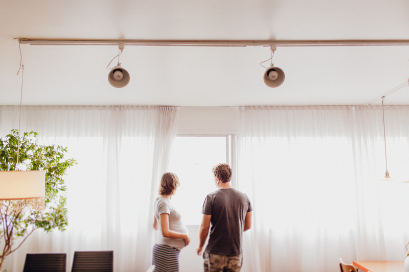Foto do Evandro e da Tetê no apartamento onde vivem. Foto do casal esperando seu primeiro filho. Foto do casal na janela feita com luz suave. Uma foto leve e espontânea. Fotografia de família, ensaio gestante, fotos de grávidas. 