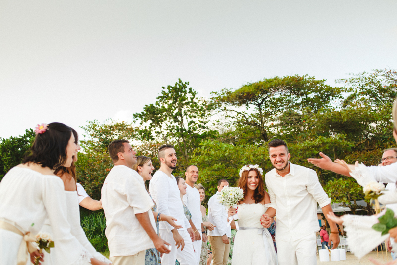 Noivos recém - casados, foto da saída dos noivos, a Mayara e o Vaz com seus padrinhos e convidados no seu casamento na praia. Fotos por Moyra e Tiago, fotógrafos de casamento