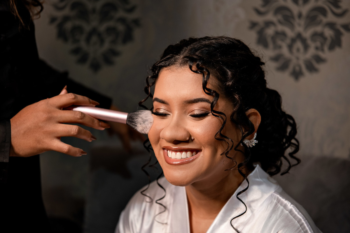 Retrato da noiva se arrumando, olhos fechados enquanto a maquiadora finaliza a produção do casamento.