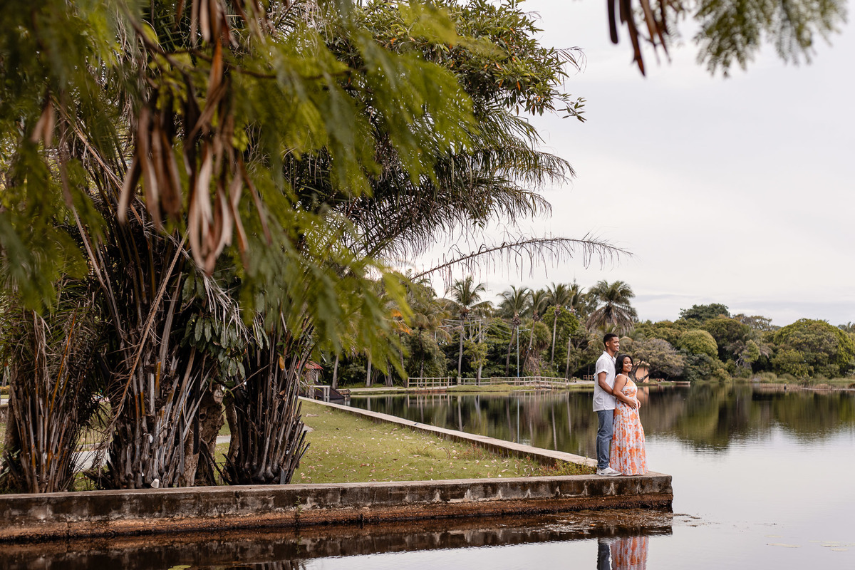 Casal em ensaio pré-nupcial na Vila de Praia do Forte — noivo abraça a noiva de vestido laranja floral à beira do lago com vegetação tropical ao fundo, fotografia por Filipe Arreciguelli