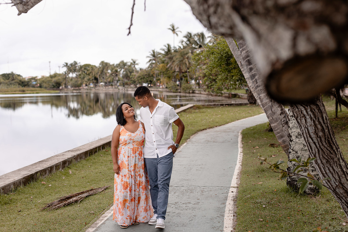 Casal apaixonado caminhando no calçadão à beira do lago na Vila de Praia do Forte — noiva com vestido floral laranja olhando para o noivo de camisa branca, ensaio pré-casamento por Filipe Arreciguelli