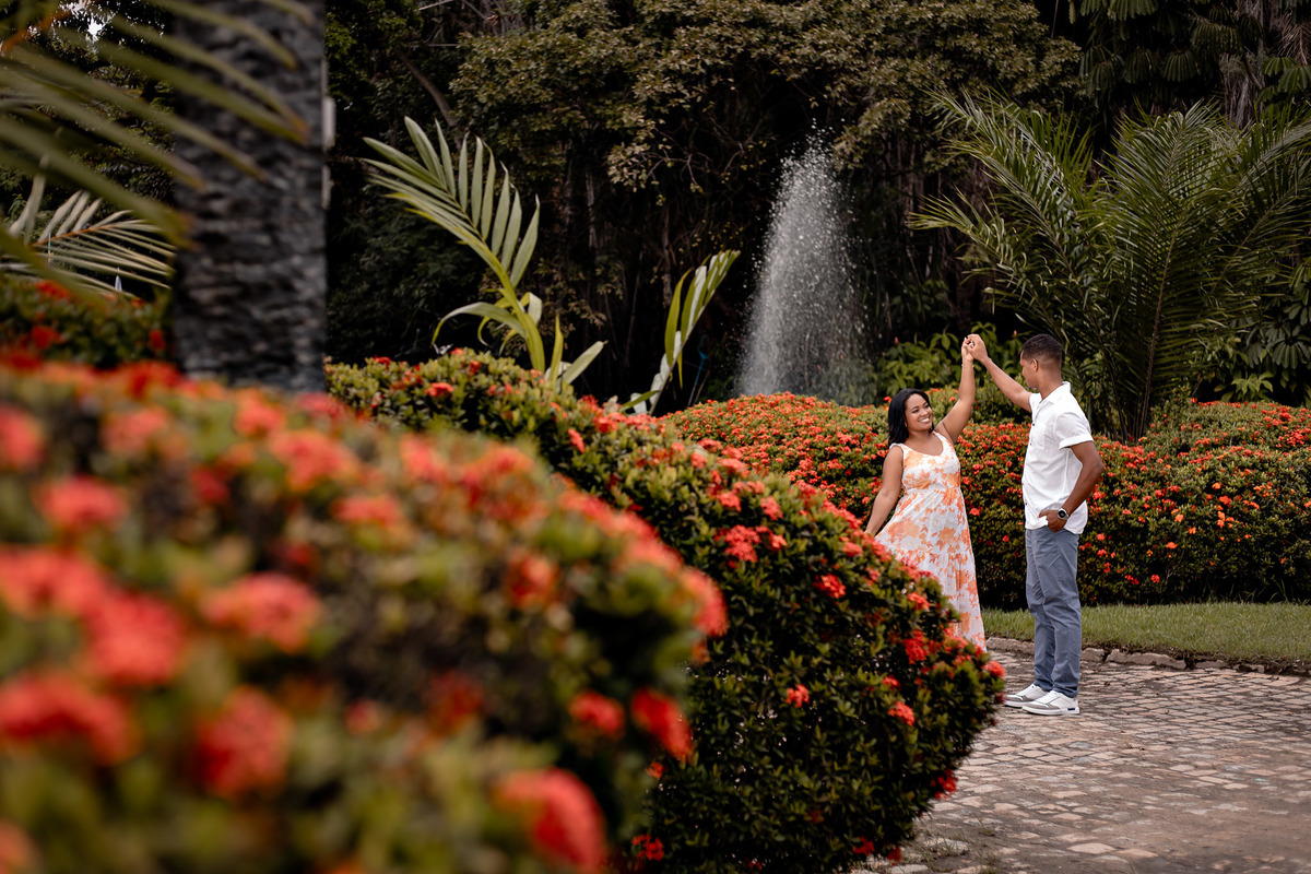 Casal dançando em jardim florido na Vila de Praia do Forte — noivo gira a noiva de vestido floral laranja entre arbustos com flores vermelhas e fonte ao fundo, ensaio pré-casamento por Filipe Arreciguelli