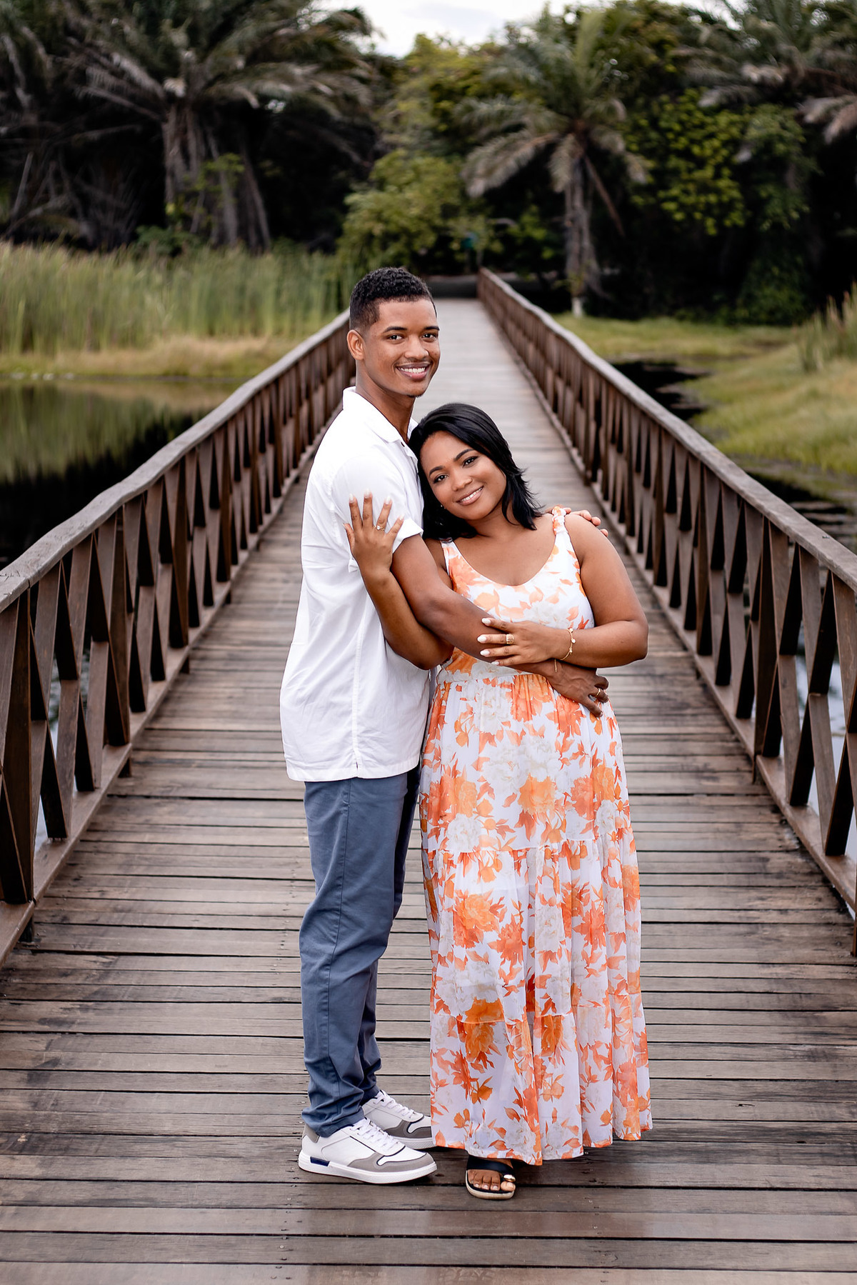 Casal abraçado na ponte de madeira da Praia do Forte — noivo de camisa branca e noiva com vestido floral laranja sorrindo para a câmera com perspectiva da ponte e coqueiros ao fundo, ensaio pré-casamento por Filipe Arreciguelli