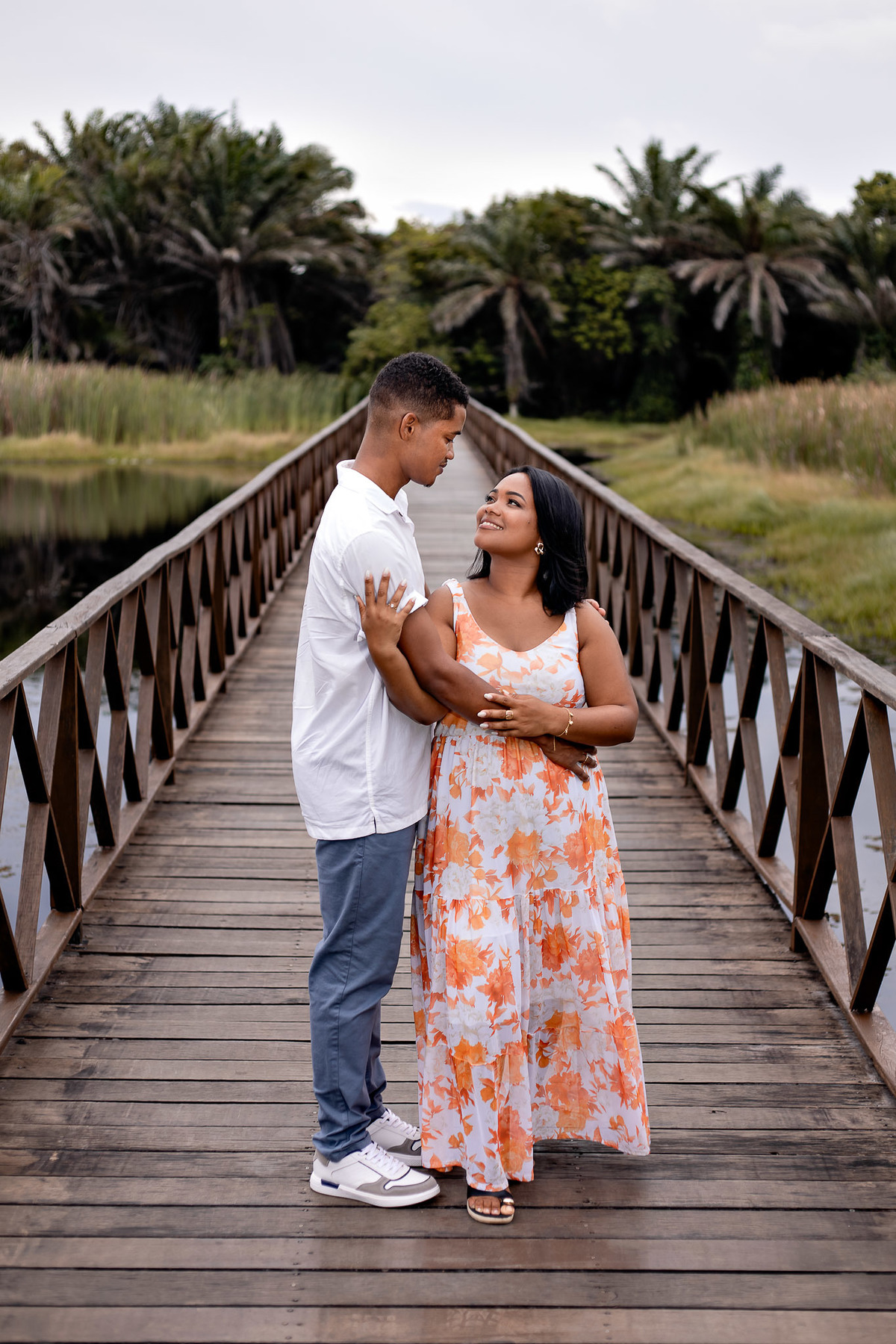 Casal apaixonado na ponte de madeira da Praia do Forte — noiva de vestido floral laranja sorrindo para o noivo de camisa branca, ensaio pré-casamento por Filipe Arreciguelli Salvador